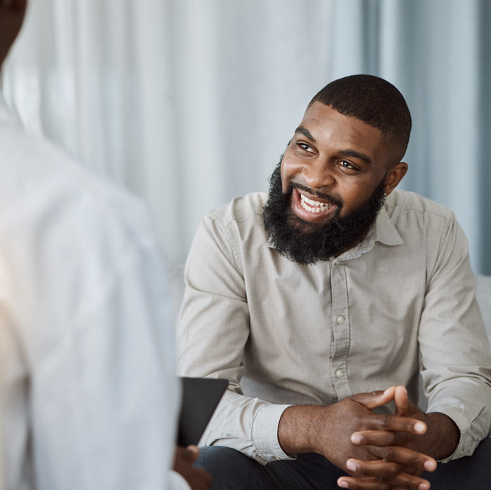 A man with a beard smiling while engaged in conversation, dressed in a light-colored shirt, against a soft background.