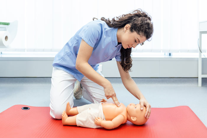 A woman in a blue shirt performing CPR on a baby doll on a red mat in a well-lit training environment.