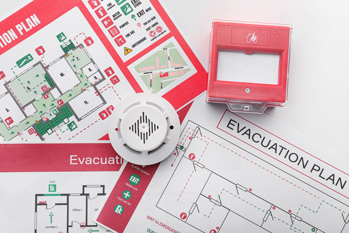 Fire alarm devices and evacuation plans displayed on a surface, featuring a smoke detector, fire alarm pull station, and detailed evacuation routes.