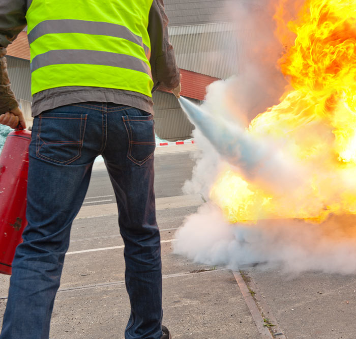 A person wearing a high-visibility vest uses a fire extinguisher to put out a fire, with smoke and flames visible.