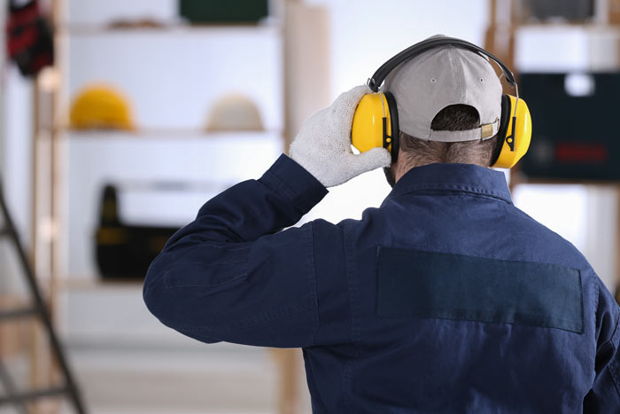 A worker wearing yellow ear protection and gloves, looking away while standing in a workspace with shelves and tools in the background.