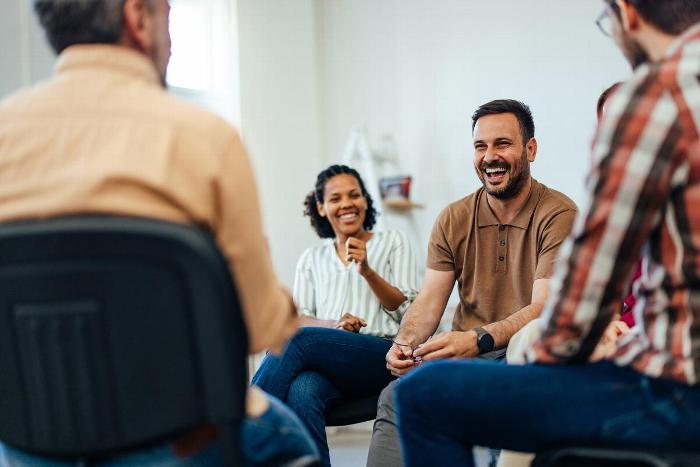 Group of diverse adults sitting in a casual setting, engaging in a friendly conversation and laughing together, showcasing community and connection.